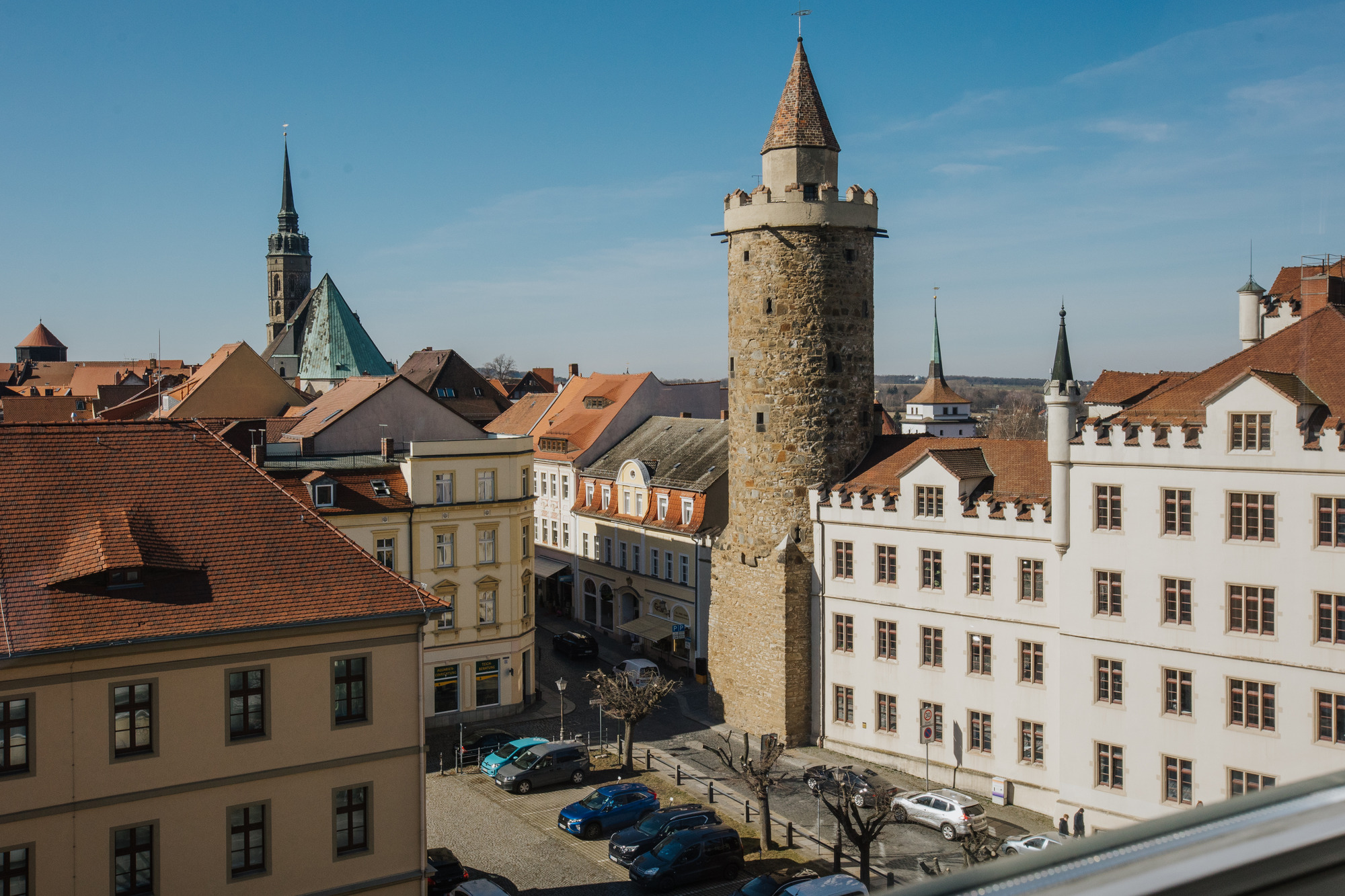 Historischer Stadtblick mit rundem Steinturm, roten Dächern, weißer Architektur und grünem Spitzturm im Hintergrund bei klarem Himmel.