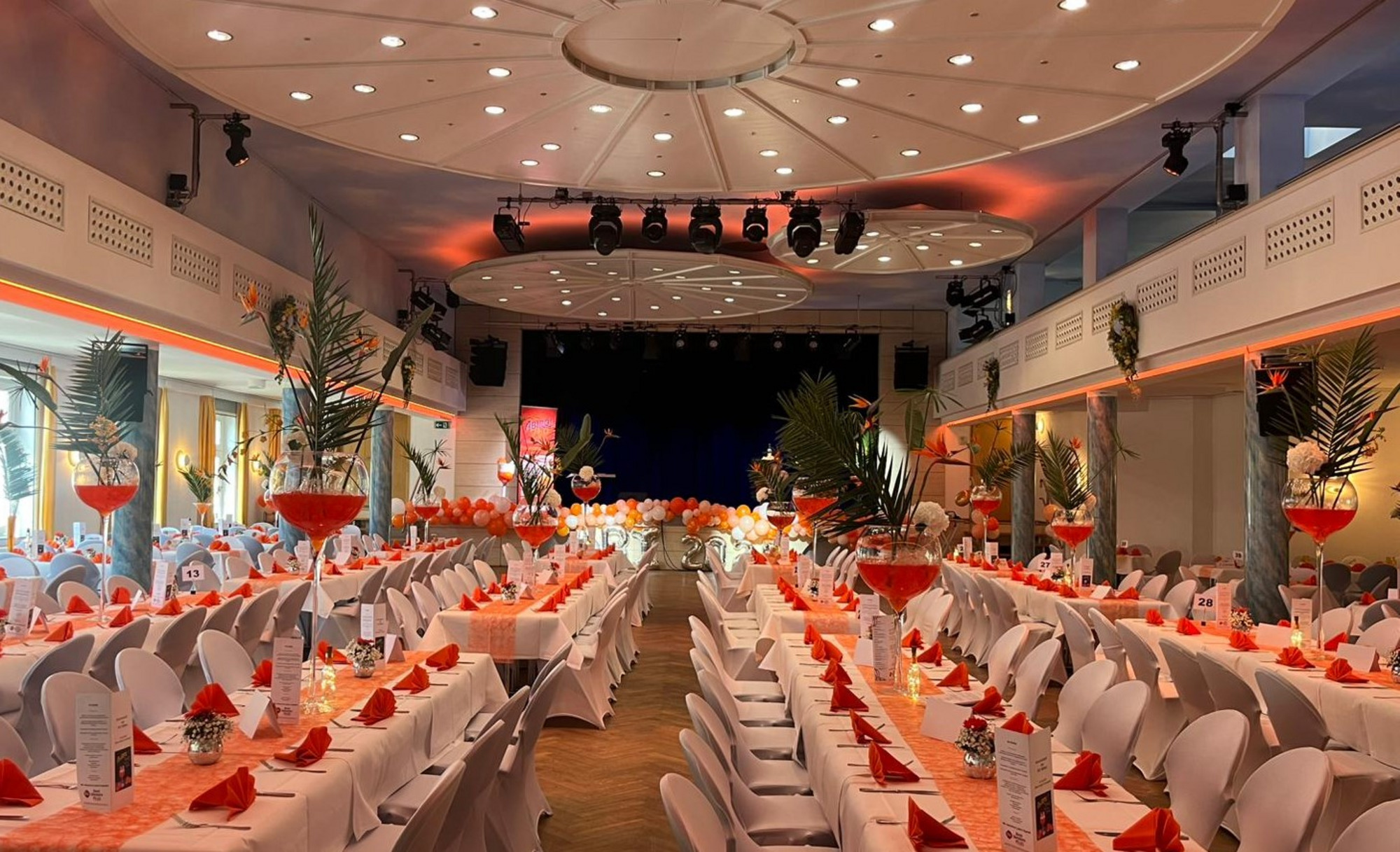 Eleganter Bankettsaal mit langen, weiß gedeckten Tischen, orangefarbenen Servietten, roten Vasen, Palmenblättern und Bühne.