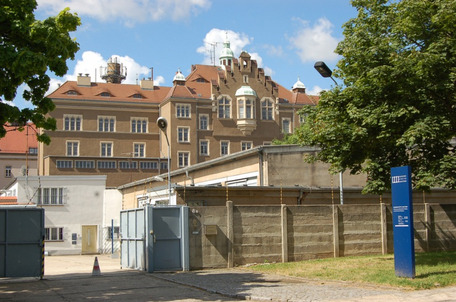Eingang zur Gedenkstätte Bautzen mit offenem Tor, blauem Hinweisschild, großem Gebäude mit roten Dachziegeln und Bäumen.
