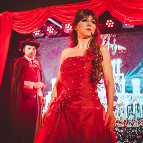 Zwei Personen in festlicher Kleidung auf Bühne mit roten Vorhängen und Scheinwerfern – Musical Night mit Abendkleid und Kostüm.